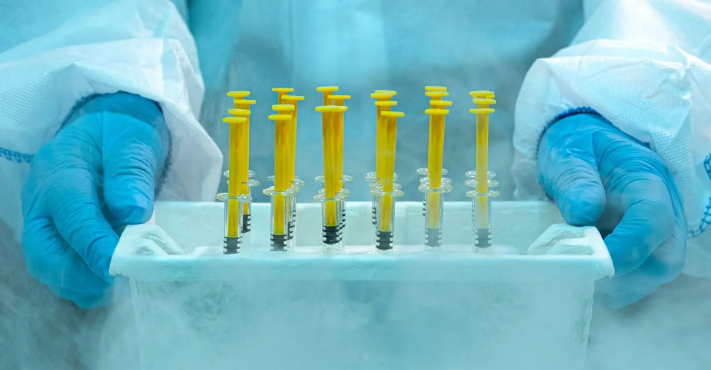 Wetenschapper draagt handschoenen en een beschermend pak, en hanteert bevroren medische injectiespuiten in een koelcontainer met zichtbaar ijs en damp.